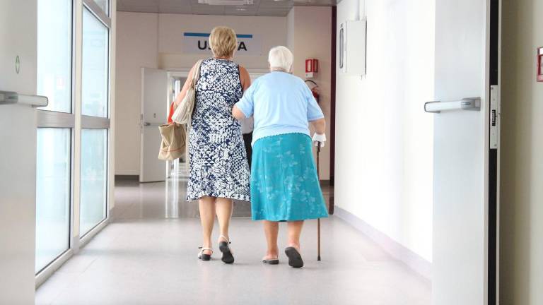 Adulta mayor camina junto a mujer más joven en un hospital.