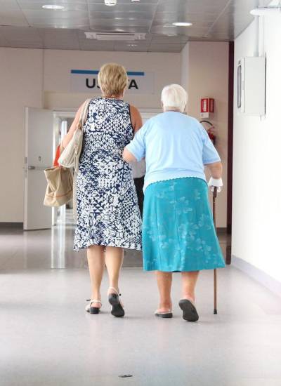 Adulta mayor camina junto a mujer más joven en un hospital.